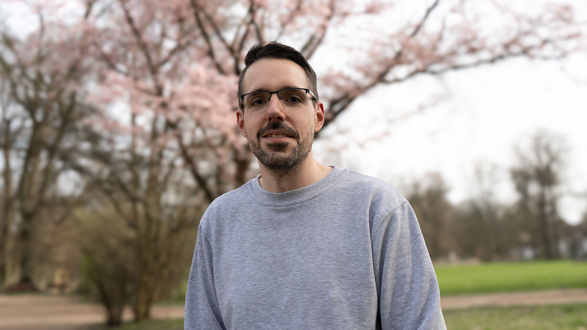 Ein Mann mit dunklen Haaren, Dreitagebart, Brille und grauem Pulli steht vor einem rosa blühenden Baum.
