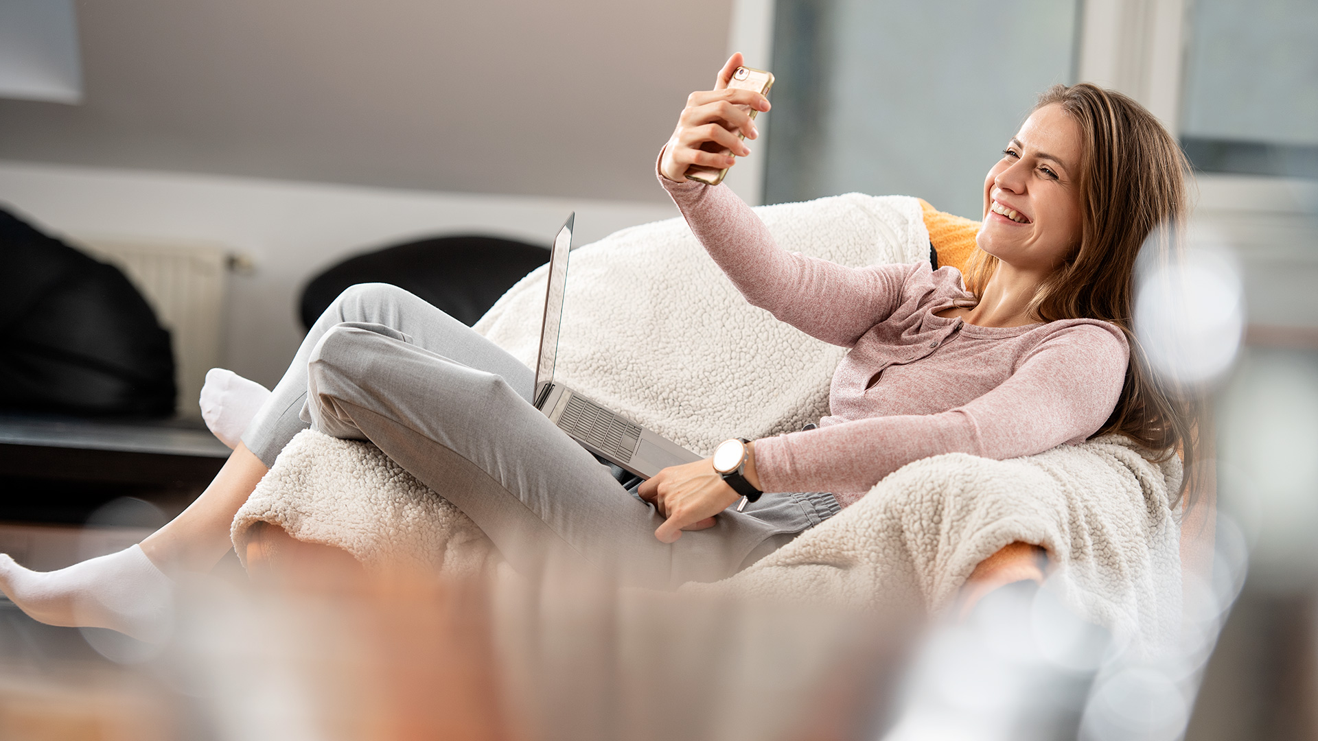 Smiling woman with a prosthetic leg relaxing in an armchair, taking a selfie with her smartphone and a laptop on her lap.