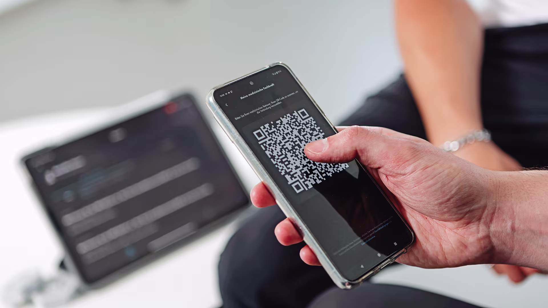 Auf dem Bild sieht man eine Hand in der ein Handy liegt. Auf dem Handy ist ein QR Code abgebildet. Im Hintergrund steht auch ein Tablet und es sitzt jemand im Hintergrund. 