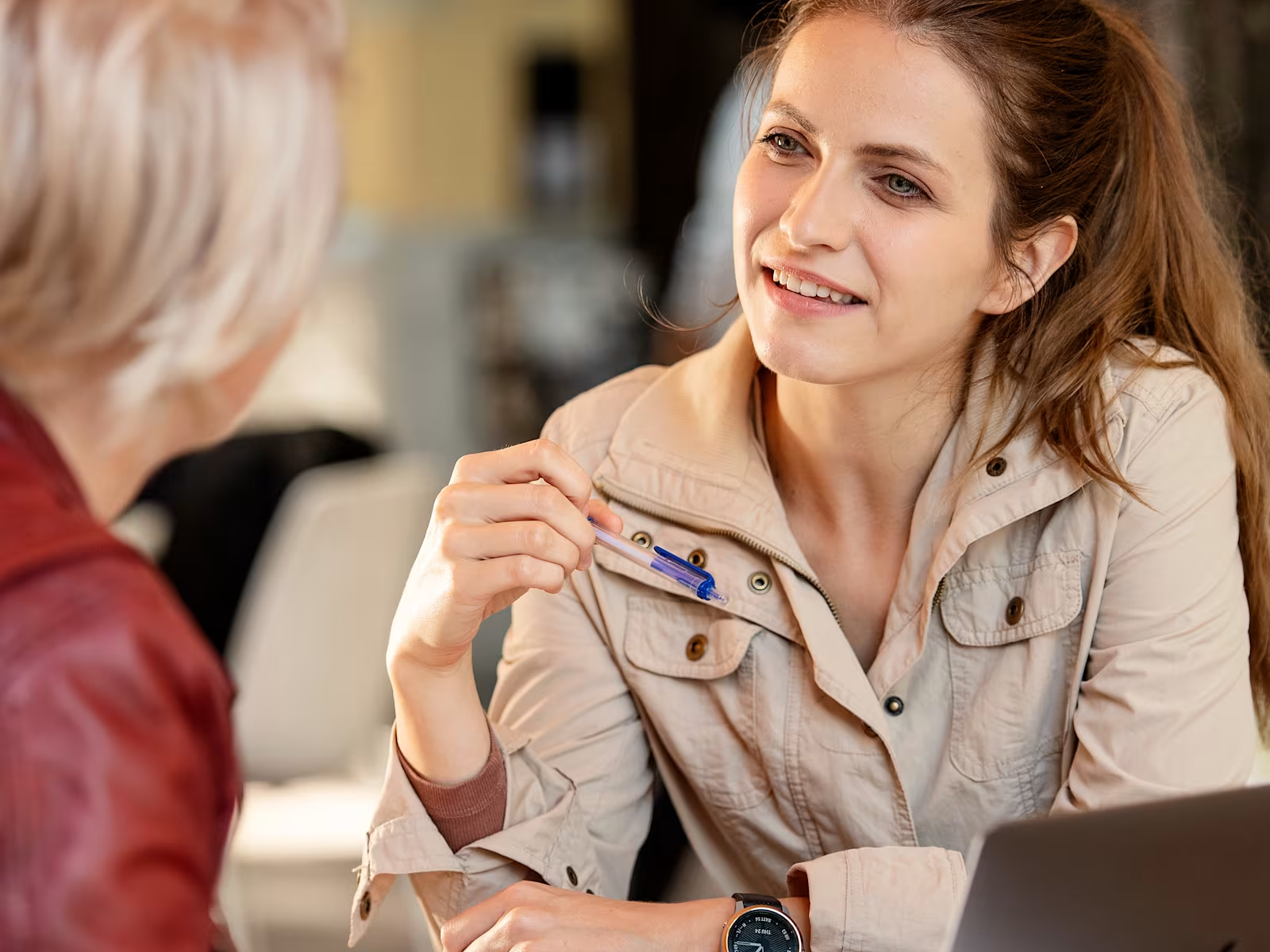 A young woman is sitting at a table in a café or work environment and is engaged in a conversation with another person. She is holding a pen and smiling attentively, with a laptop and documents placed in front of her. The scene feels calm, professional, and conversational, showing a prosthetics user in an everyday social setting.