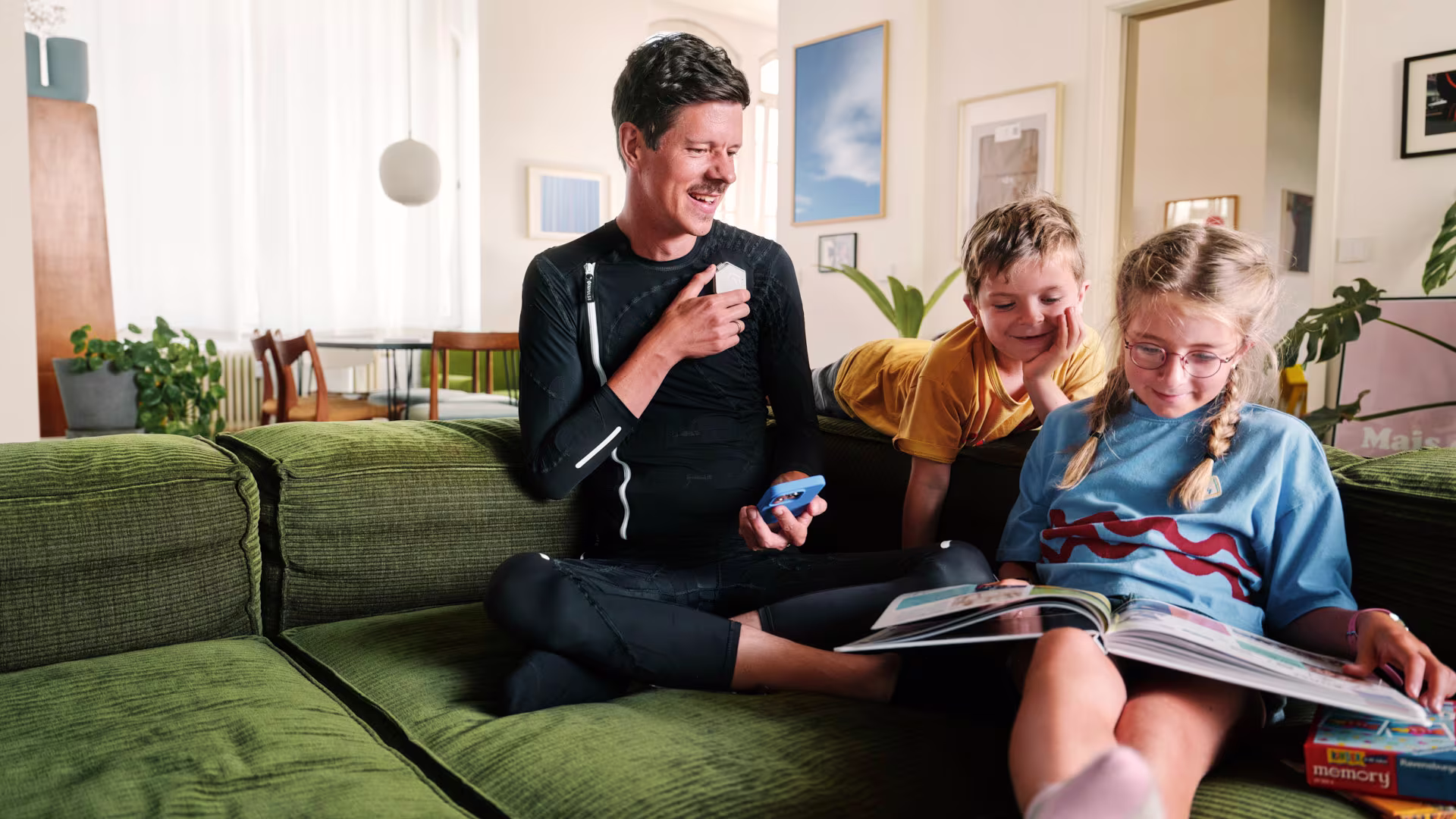 Ein Mann sitzt lächelnd auf einem grünen Sofa. Er trägt den schwarzen Ganzkörper-Neuromodulationsanzug exopulse suit von Ottobock. Er hält ein blaues Smartphone in der Hand. Neben ihm sitzen zwei Kinder, ein Junge und ein Mädchen. Das Mädchen mit Brille und zwei Zöpfen liest in einem großen Buch, während der Junge über ihre Schulter schaut. Sie befinden sich in einem gemütlichen Wohnzimmer mit Pflanzen und moderner Einrichtung im Hintergrund.