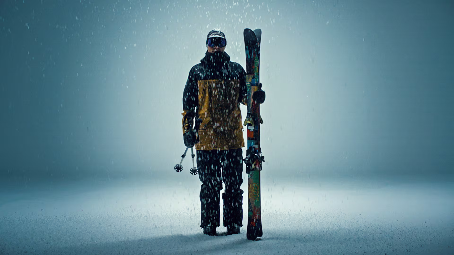 Ein Skifahrer in voller Skibekleidung hält die Skier in seiner linken Hand und steht vor einem dunklen Hintergrund.