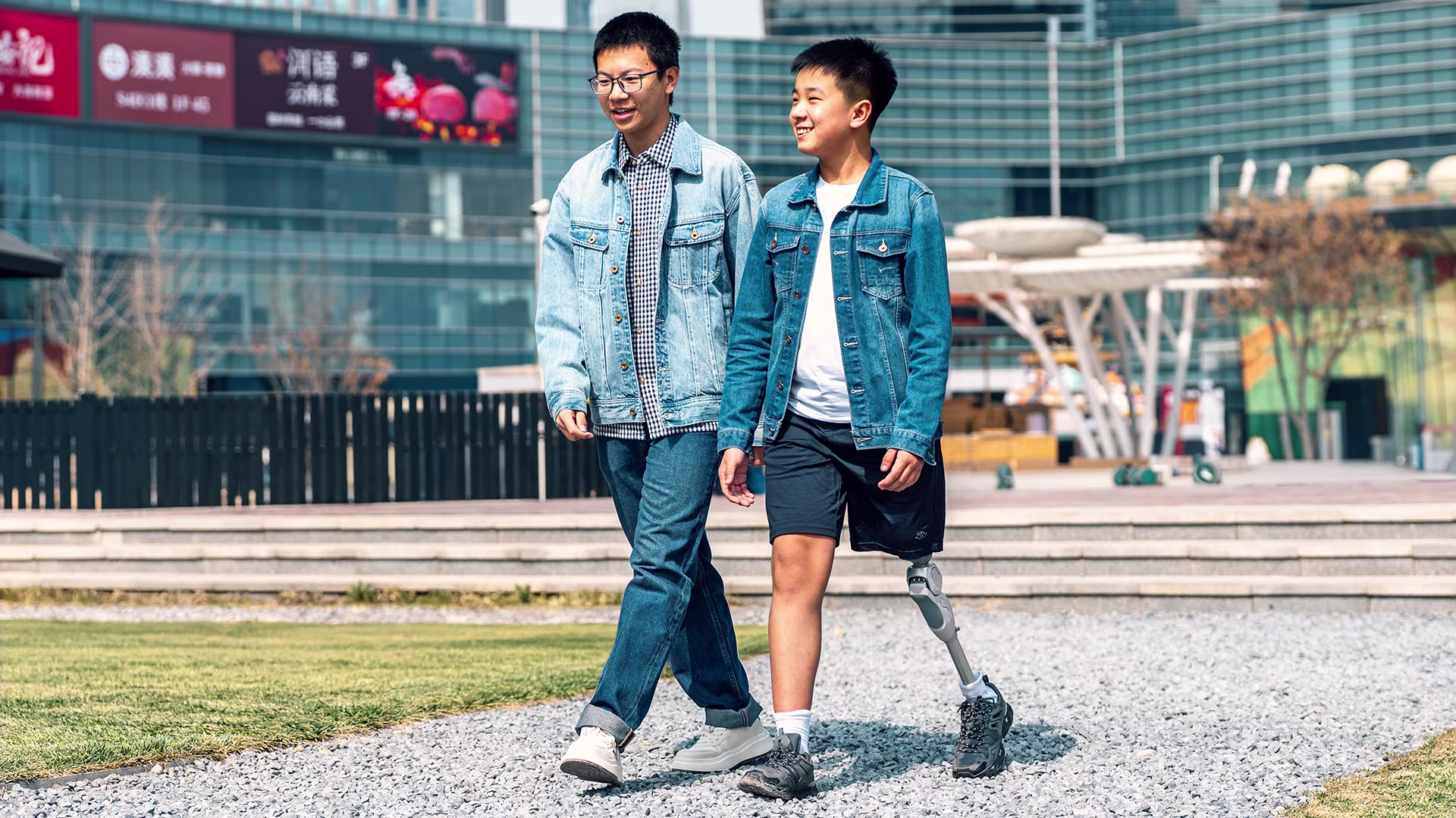 Two teenage boys walk side by side in a mall courtyard; one has a prosthetic leg.