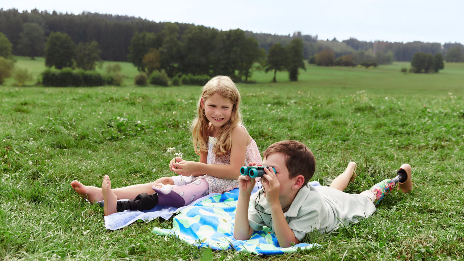Two children with Ottobock leg prostheses sitting together on a grass lawn outdoors.