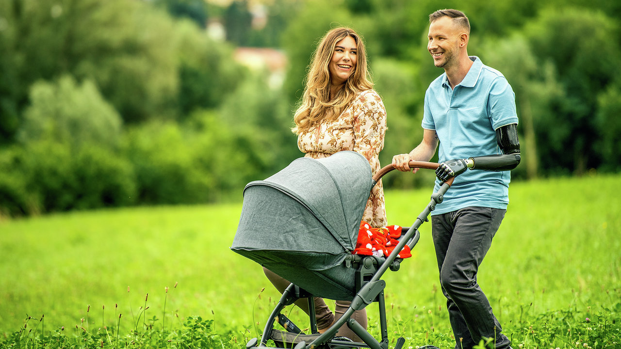 Ein Armprothesenträger spaziert mit seiner Frau und seinem Kind in einem Kinderwagen. Er trägt das DynamicArm-Ellbogengelenk und die bebionic Hand von Ottobock.