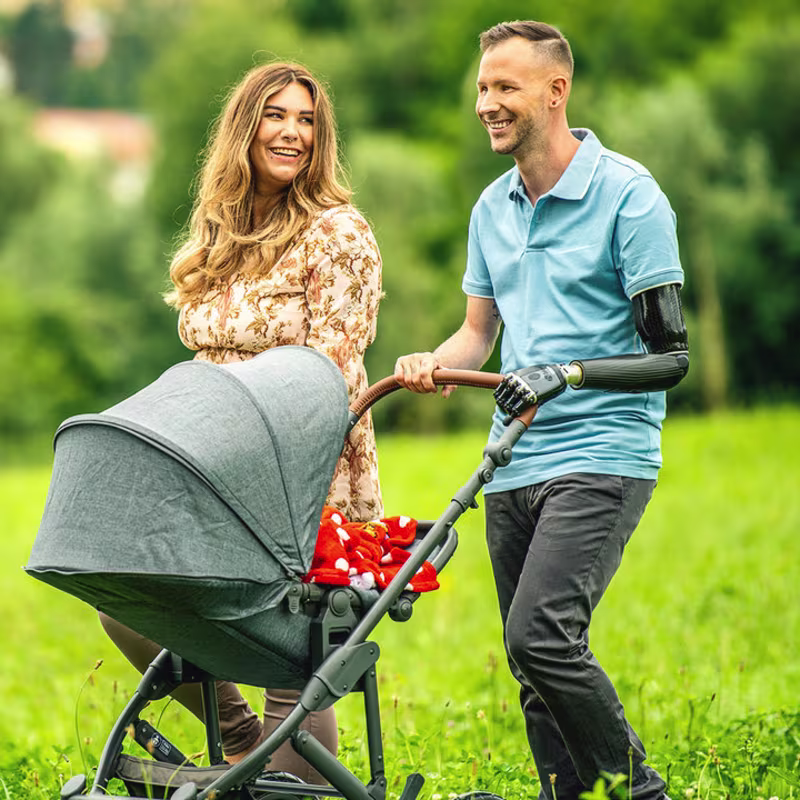 Ein Armprothesenträger spaziert mit seiner Frau und seinem Kind in einem Kinderwagen. Er trägt das DynamicArm-Ellbogengelenk und die bebionic Hand von Ottobock.