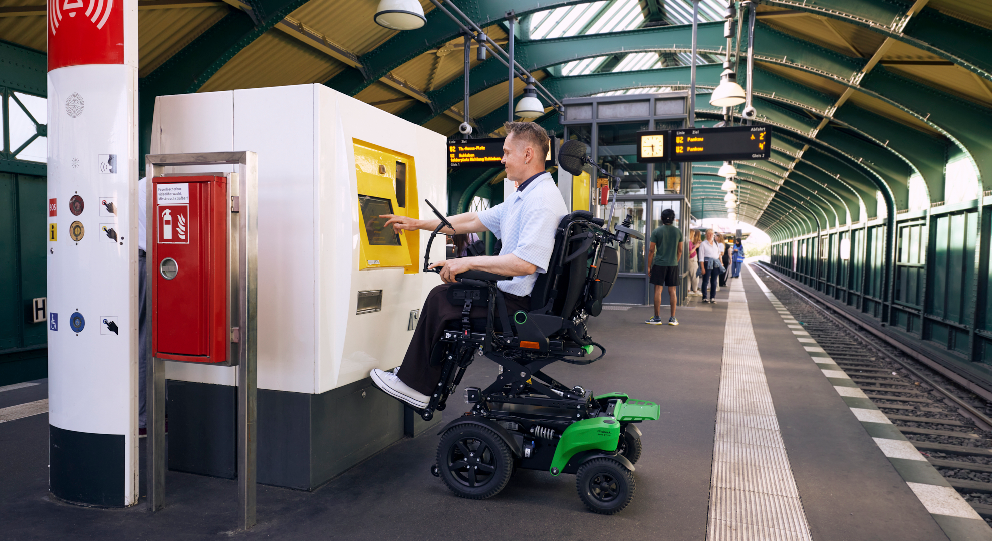 Un homme en fauteuil roulant électrique utilise un distributeur automatique de billets à écran tactile sur le quai d'une gare accessible.