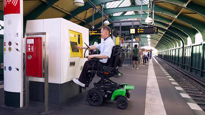 Erişilebilir bir tren istasyonu peronunda, elektrikli tekerlekli sandalye kullanan bir adam dokunmatik bilet makinesini kullanıyor.