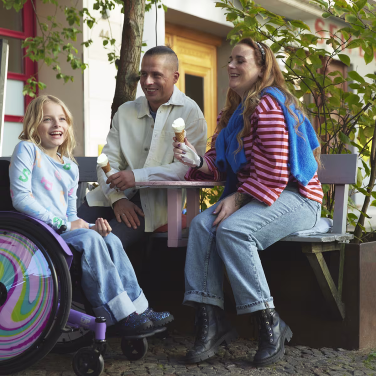 A young girl, Nomine, in her kidevo wheelchair sits outside with her parents as they all laugh while enjoying vanilla ice cream cones.