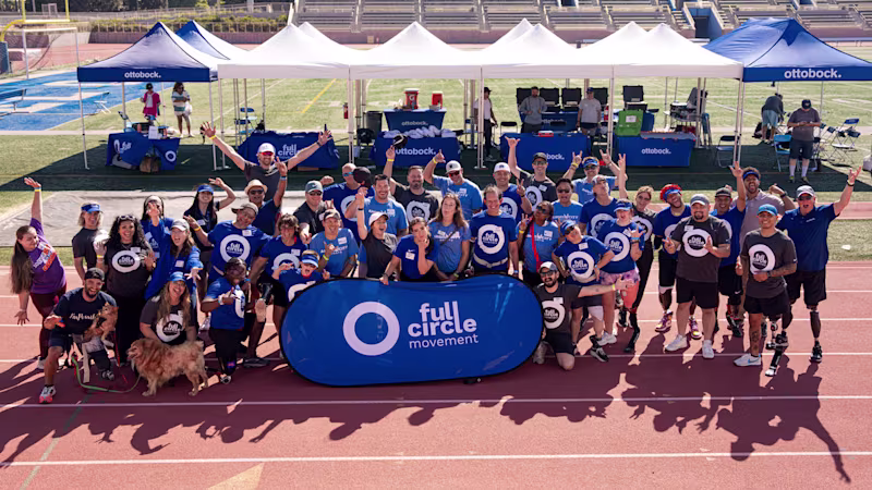 A large group of people standing on a running track on a sunny day gathered around a blue sign that says Full Circle Movement