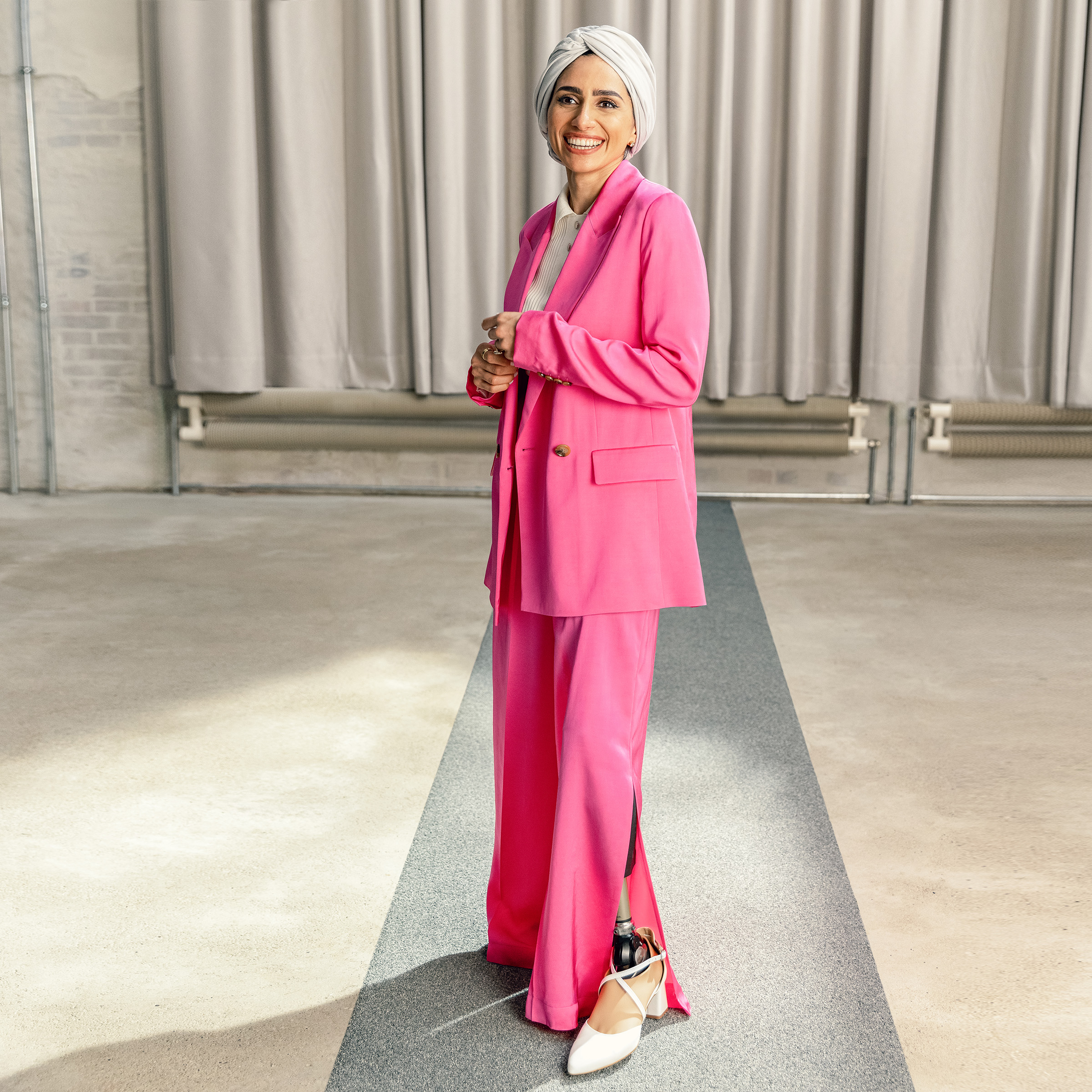 Protez ayak kullanıcısı Zainab gülümsüyor ve pembe bir takım elbise ve Ottobock Taleo Adjust ile yüksek topuklu beyaz ayakkabılar giyiyor