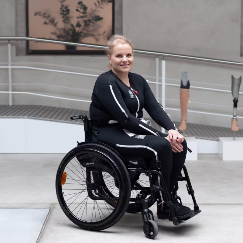 Ortopedi kliniği ortamında Exopulse Mollii Elbisesi giyen, Ottobock Avantgarde tekerlekli sandalyede gülümseyen bir kadın.