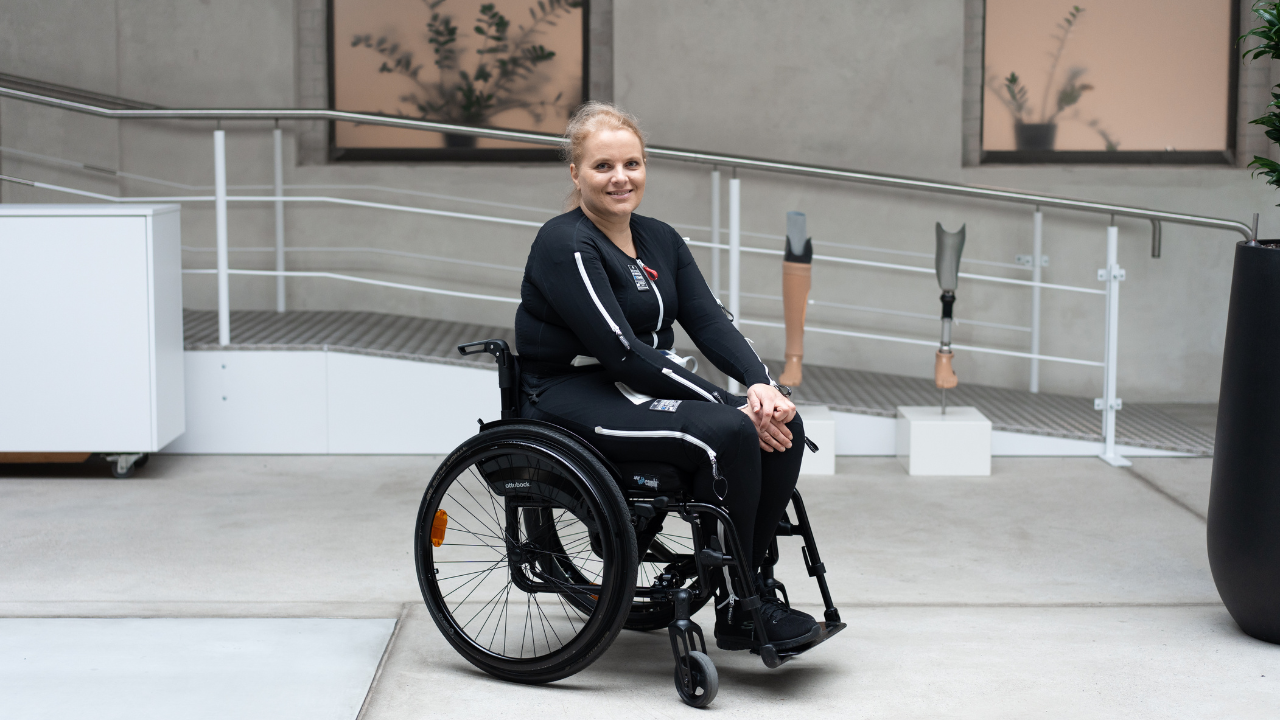 A smiling woman in an Ottobock Avantgarde wheelchair, wearing the Exopulse Mollii Suit in an orthopedic clinic setting.