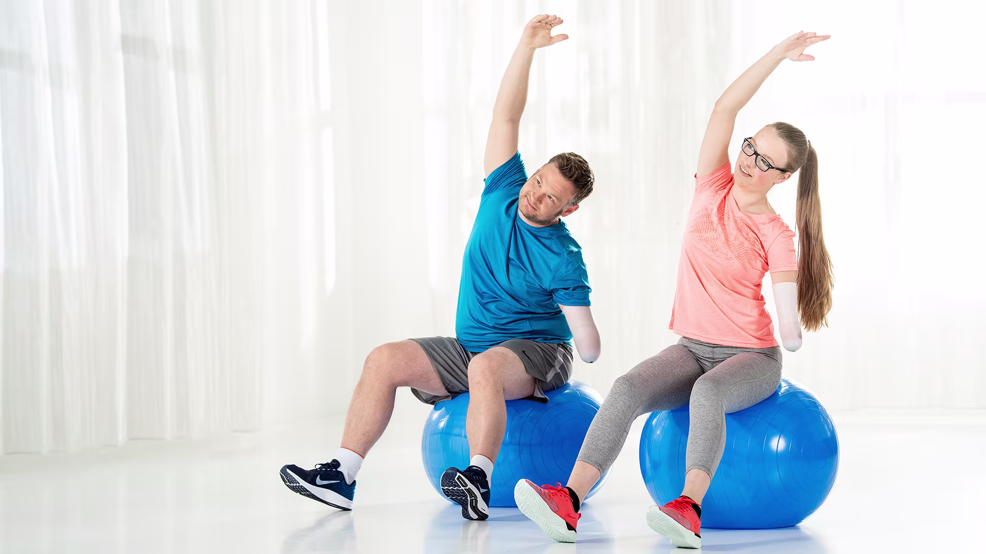 A woman and a man with an arm amputation sit next to each other on exercise balls and stretch their right arm up over their heads