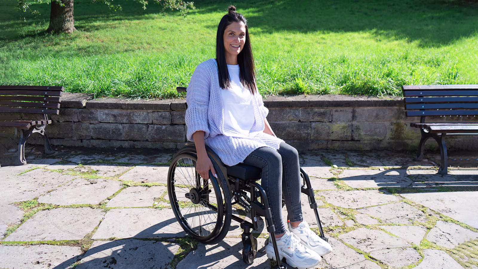 Eine weiblich gelesene Person mit dunkelen langen Haaren sitzt im Motus Aktivrollstuhl von Ottobock. Sie lächelt in die Kamera. Im Hintergrund erkennt man eine grüne Wiese, eine Parkmauer und zwei braune Sitzbänke.