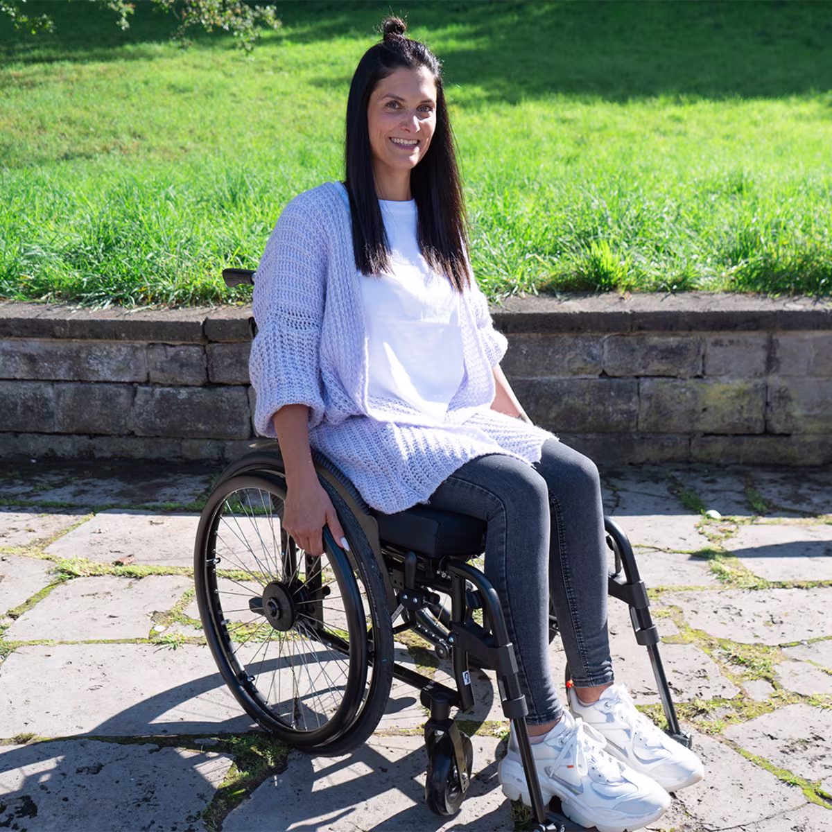 A smiling woman with brown hair and a white top sits in an Ottobock wheelchair on a terrace in front of a meadow