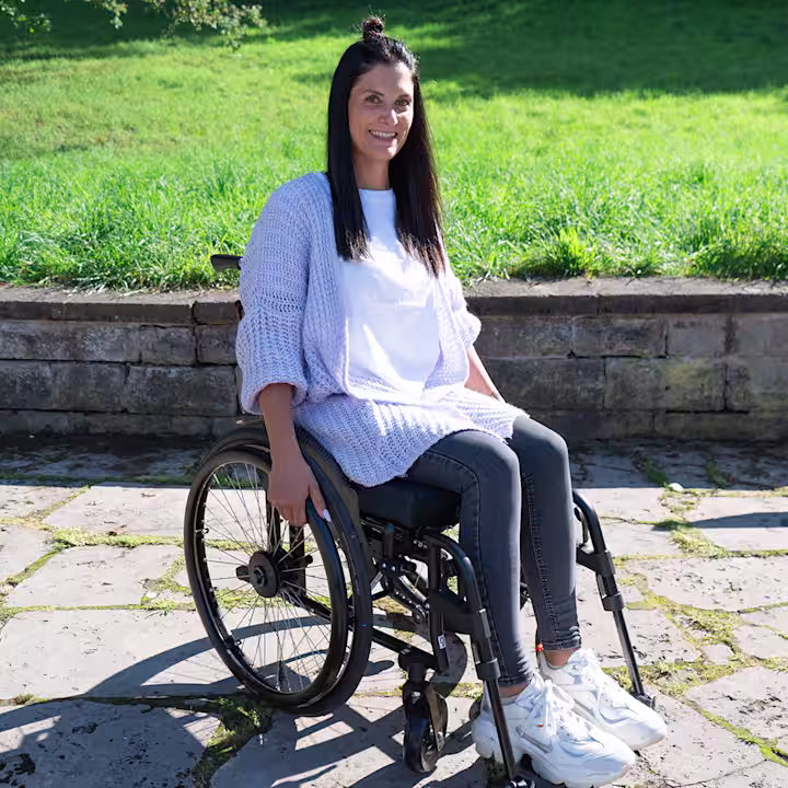 A smiling woman with brown hair and a white top sits in an Ottobock wheelchair on a terrace in front of a meadow
