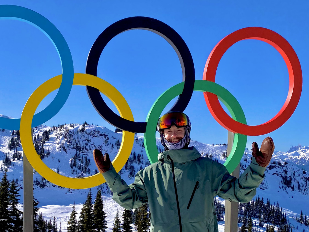 Man in ski gear standing happily in front of a statue of the Olympic rings with arms stretched wide 