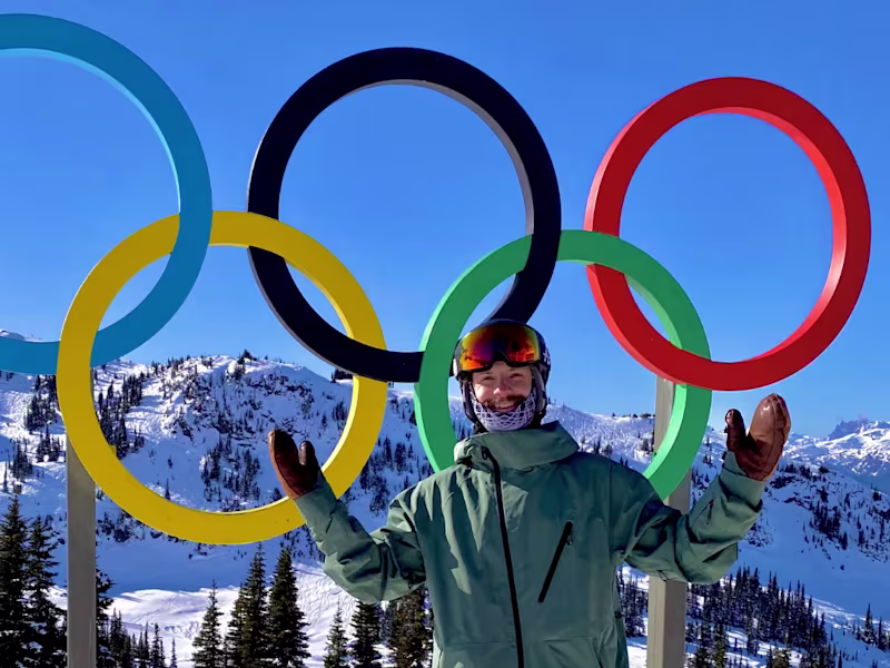Man in ski gear standing happily in front of a statue of the Olympic rings with arms stretched wide