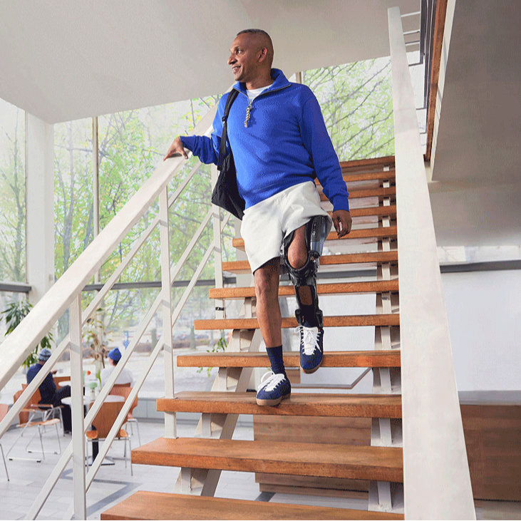 A man walks down stairs in a brightly lit cafe while smiling and looking wistfully out the window. He is wearing an Ottobock C-Brace orthotic brace