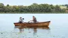 A man with a partial silicone hand prosthetic paddles a wooden canoe in a lake with a woman and dog companion