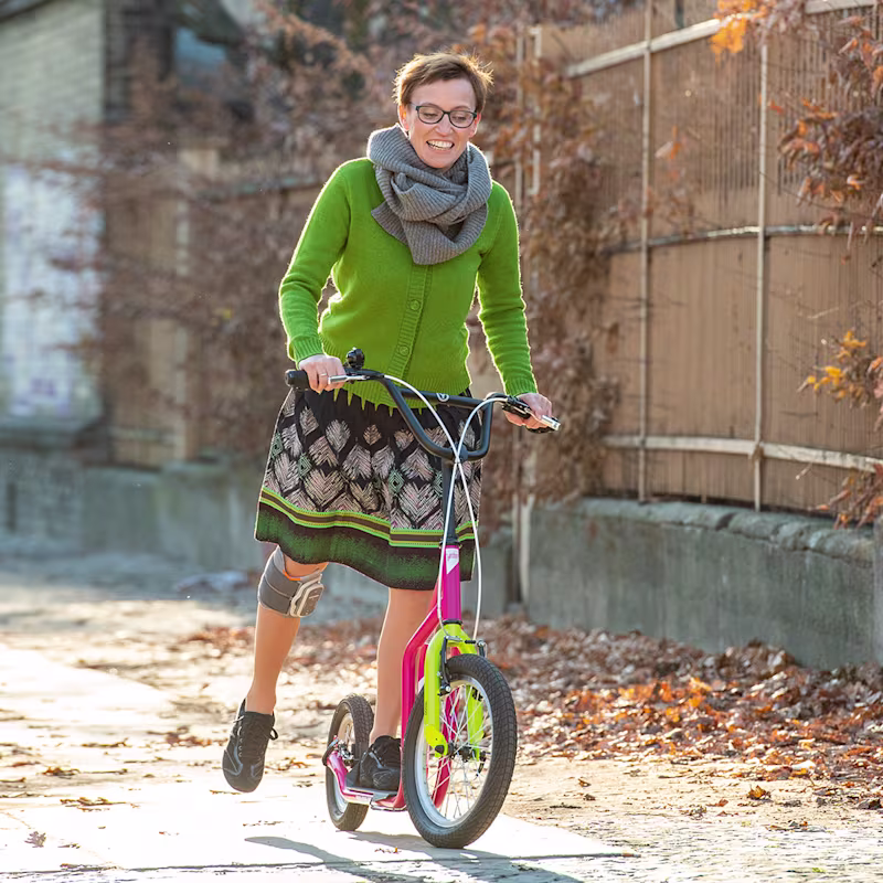 A person read as a woman is wearing Ottobock’s L100 Go L100 Go on her right leg. She stands on a colourful scooter, smiles, and uses her right leg to swing the ball.