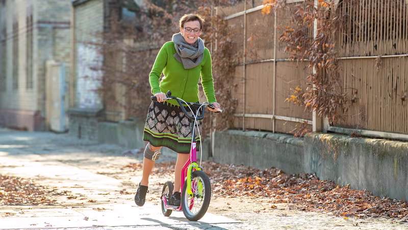 Eine weiblich gelesene Person trägt am rechten Bein das L100 Go von Ottobock. Sie steht auf einem bunten Roller, lächelt und gibt mit dem rechten Bein Anschwung.