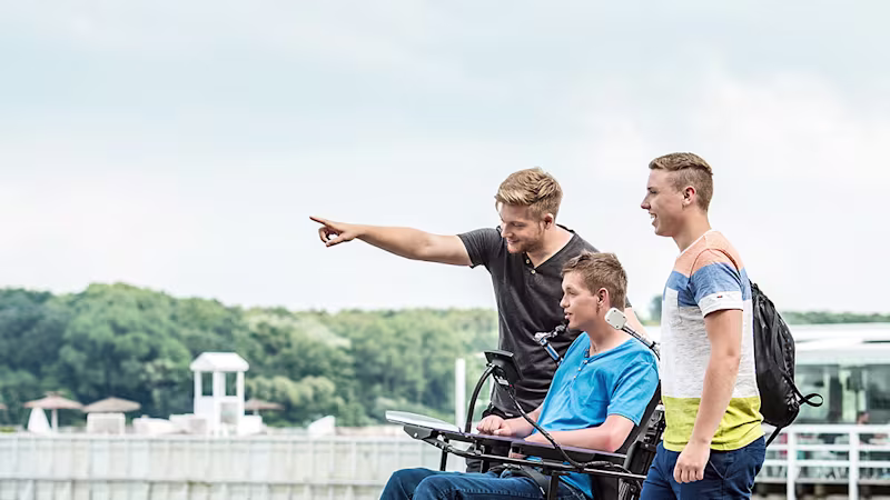 Drei junge Männer, einer davon im Rollstuhl, stehen an einem See und beobachten etwas in der Ferne.