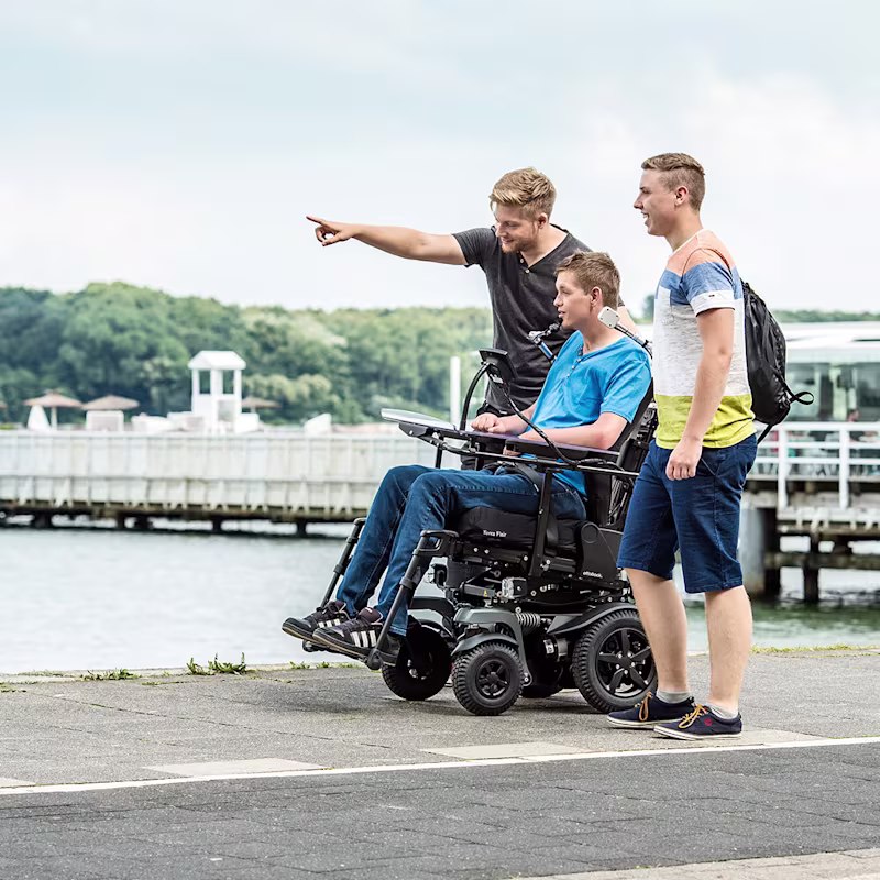 Drei junge Männer, einer davon im Rollstuhl, stehen an einem See und beobachten etwas in der Ferne.