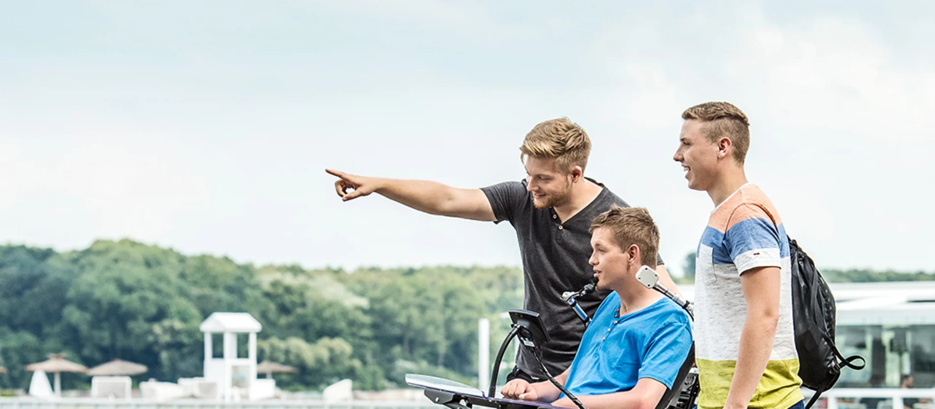 Drei junge Männer, einer davon im Rollstuhl, stehen an einem See und beobachten etwas in der Ferne.