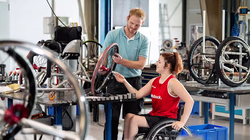 Ein Techniker steht mit einer Anwenderin in einem Rollstuhl in einer Werkstatt und reparieren ein Rad eines Rollstuhls.
