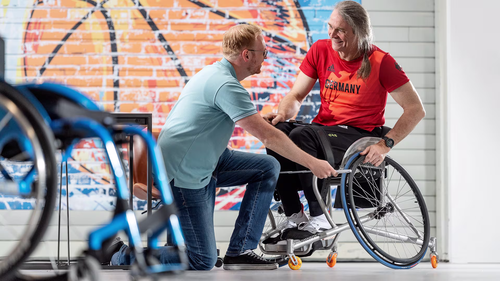 Ein Mann sitzt in einem Invader Sport Rollstuhl von Ottobock. Ein anderer Mann kniet vor dem Rollstuhl und misst mit einem Zollstock dessen Breite aus.
