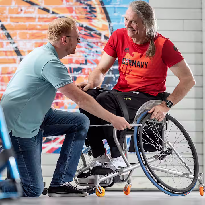Ein Mann sitzt in einem Invader Sport Rollstuhl von Ottobock. Ein anderer Mann kniet vor dem Rollstuhl und misst mit einem Zollstock dessen Breite aus.