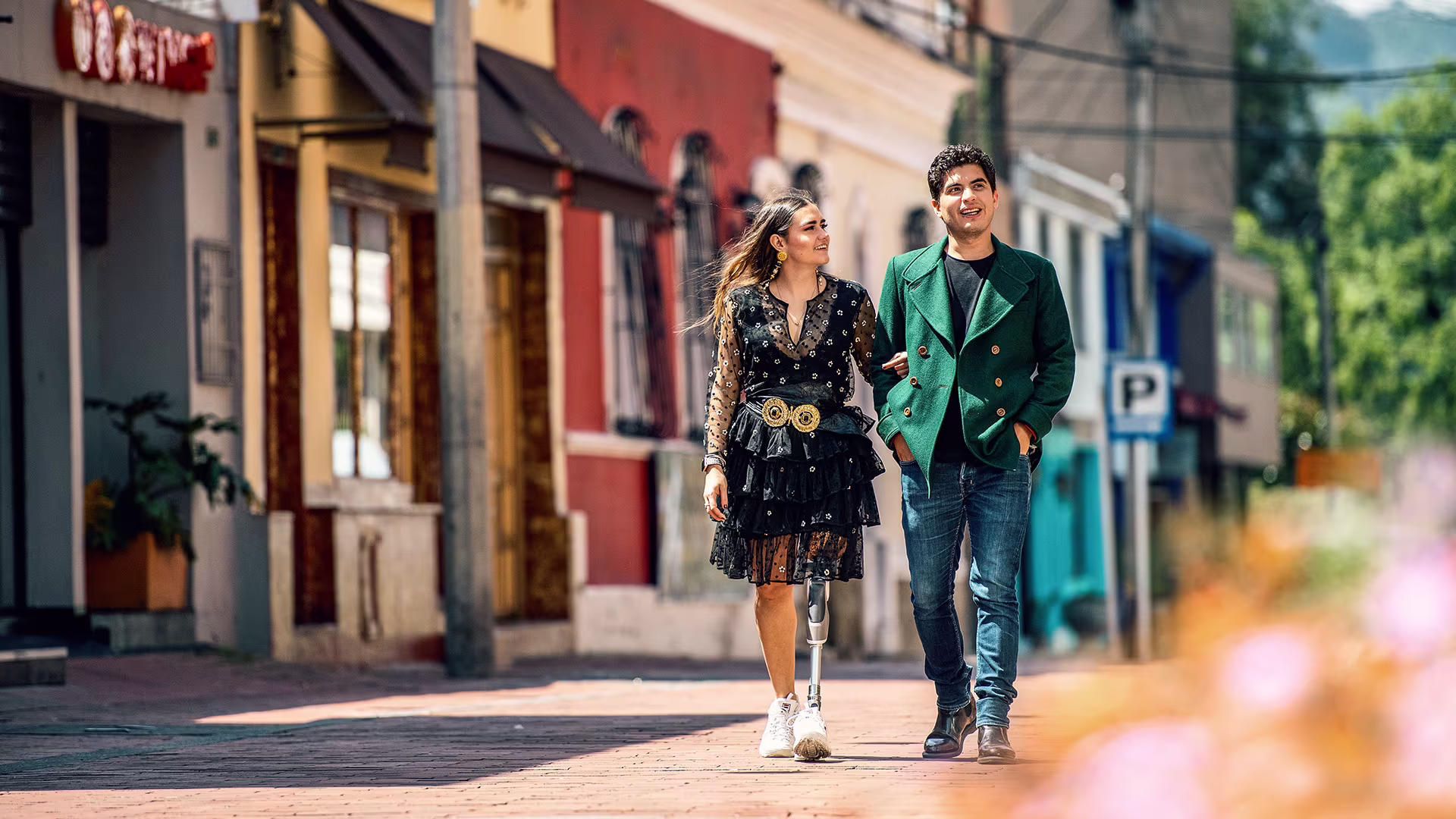 A young women with a leg prosthesis walks along a streets, linking arms with the man beside her.