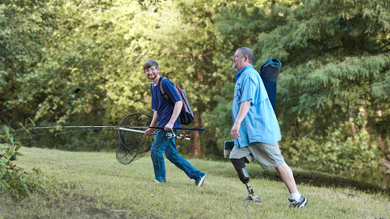 Two men transport various fishing equipment through a forest. The man in the forefront of the photo sports the custom Ottobock Taleo bionic foot on his right leg.