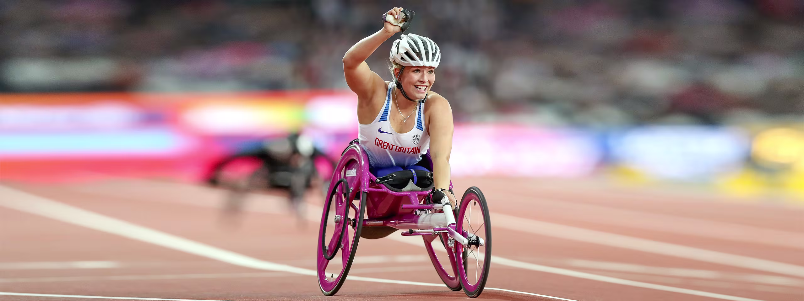 A young woman in a pink racing wheelchair and helmet on a racing track, raising her fist and smiling.
