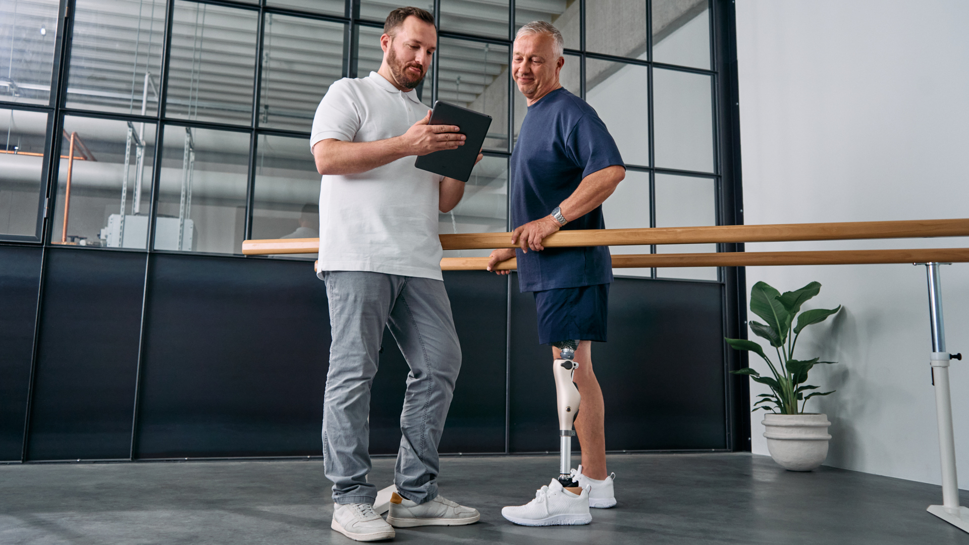 A patient stands in parallel bars wearing a microprocessor knee while a CPO shows settings on a tablet.