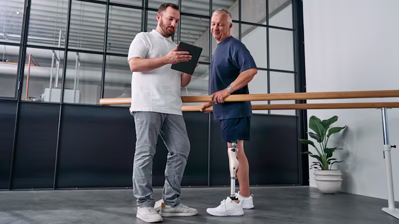 A patient stands in parallel bars wearing a microprocessor knee while a CPO shows settings on a tablet.