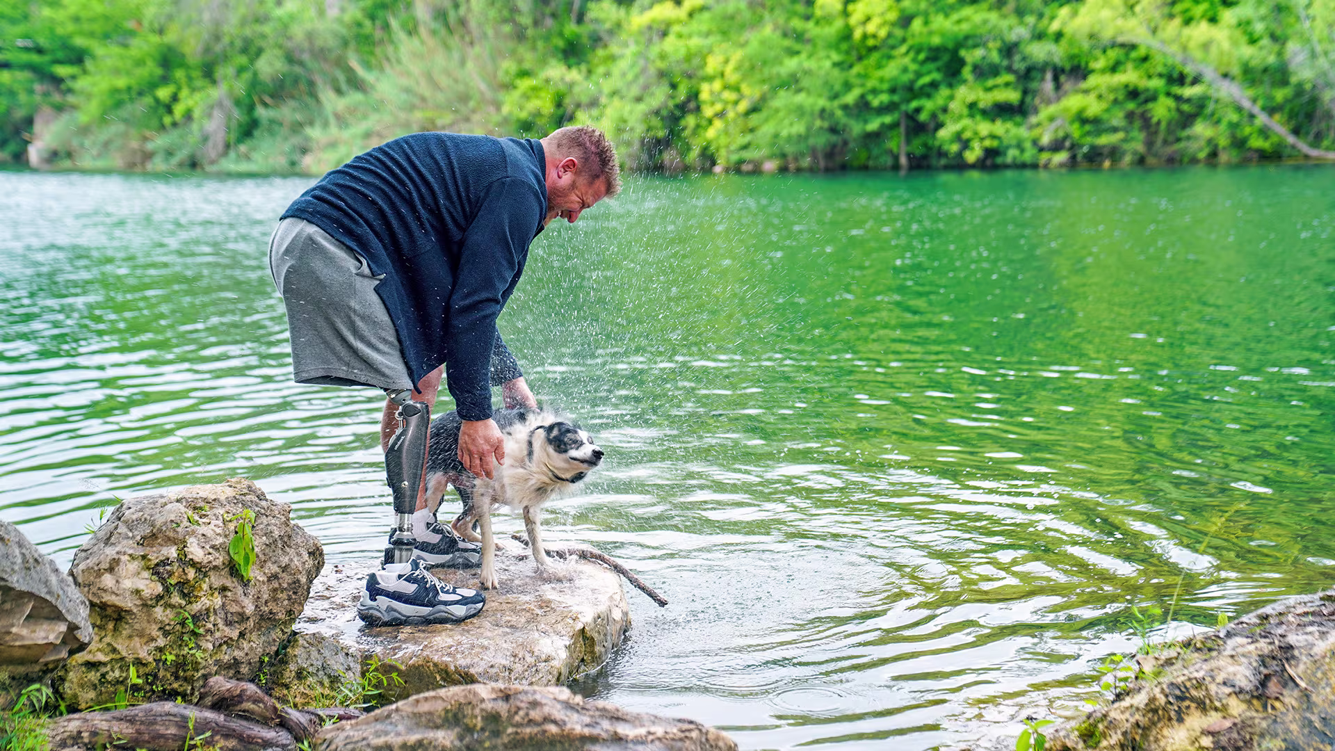 A male C-Leg user pets his wet dog close to a lake.