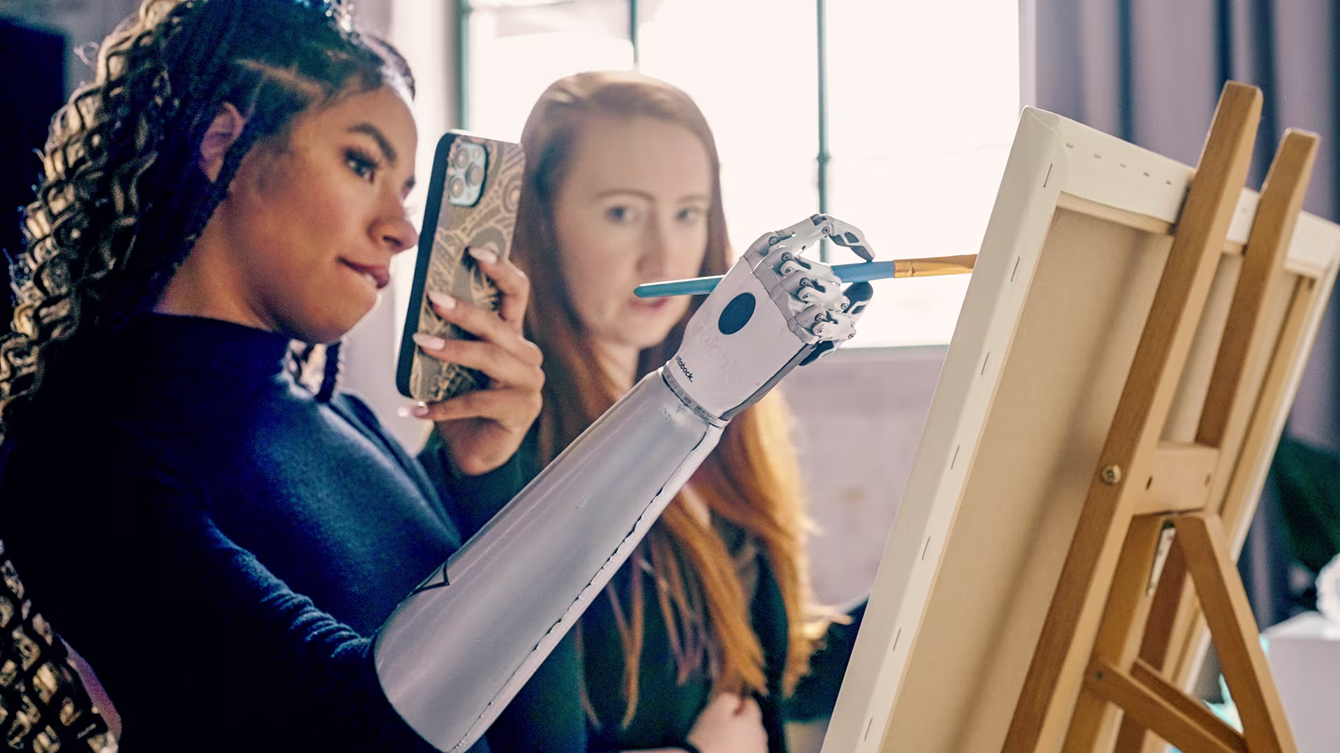 Two women in front of an easel. One is holding a pencil in her bebionic prosthetic hand and taking a selfie. The other woman is watching from the background.