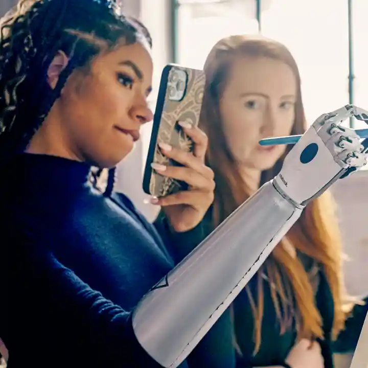 Two women in front of an easel. One is holding a pencil in her bebionic prosthetic hand and taking a selfie. The other woman is watching from the background.