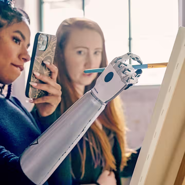 Bir şövalenin önünde iki kadın. Biri bebiyonik protez eliyle kalem tutuyor ve selfie çekiyor. Diğer kadın ise arka plandan izliyor.