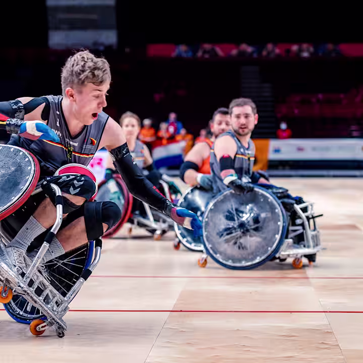 Bei einem rasanten Rollstuhl Basketballspiel fährt ein Mann auf einem Rad seines Invader Rugby. Im Hintergrund sind mehrere Sportler im Rollstuhl zu erkennen.