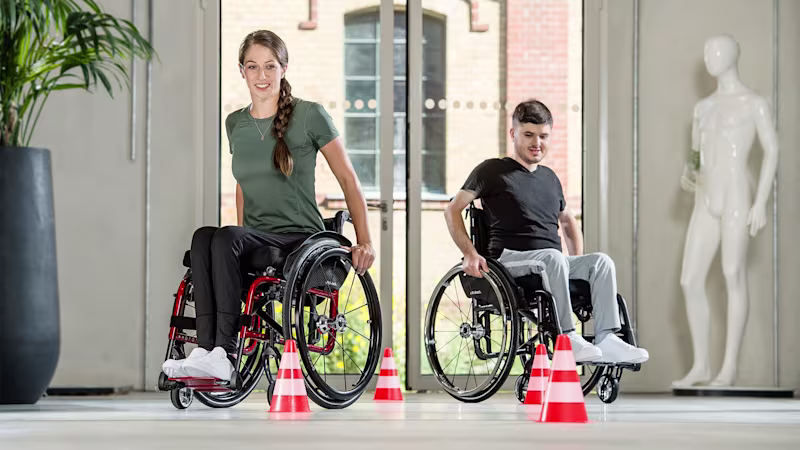 Eine weibliche und eine männliche Person sitzen im Avantgarde Aktivrollstuhl von Ottobock und umfahren im Slalom Pylonen in einem Therapieraum.