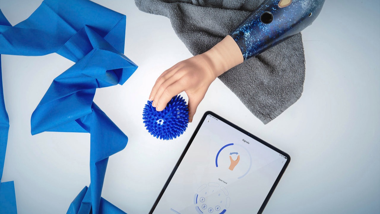 A brightly lit tabletop with an Ottobock speedhand grasping a blue ball, a blue elastic exercise band, and a tablet showing Ottobock's myosmart functionality in the connectgrip app