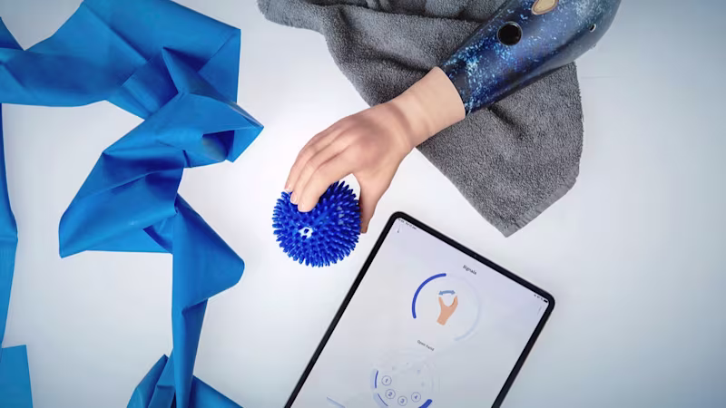A brightly lit tabletop with an Ottobock speedhand grasping a blue ball, a blue elastic exercise band, and a tablet showing Ottobock's myosmart functionality in the connectgrip app