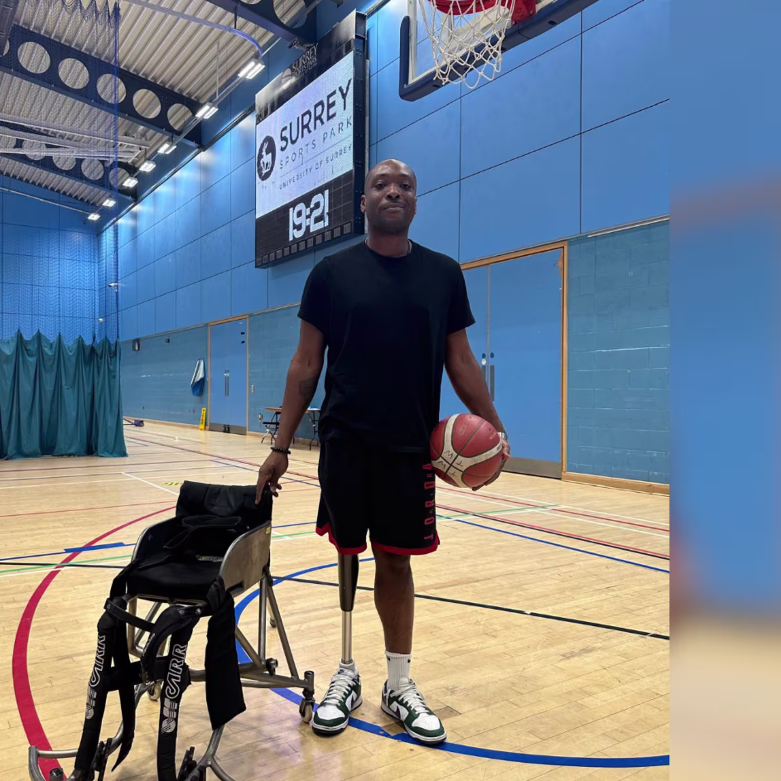 C-Leg user Vickson stands on a basket ball court with an orange basketball under one arm and holding onto his wheelchair for basketball in the other.