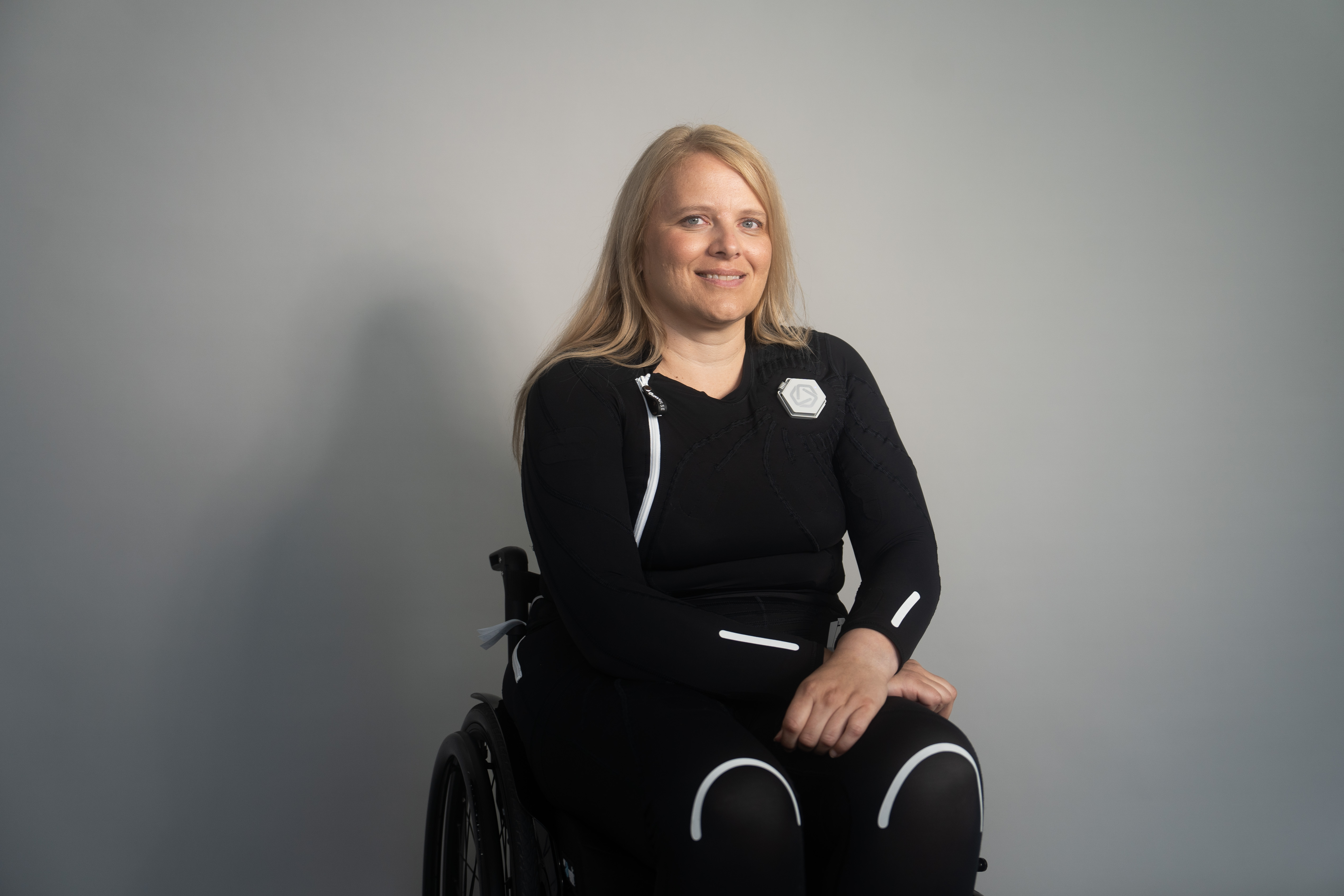 Eine lächelnde Frau in einem Avantgarde Rollstuhl von Ottobock. Sie trägt den Exopulse Mollii Suit. Im Hintergrund ist eine orthopädischen Klinik.