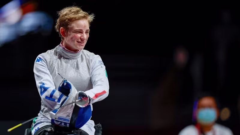 Young woman with short hair wearing a grey and blue full-body suit in a wheelchair, holding a fencing sword and smiling.