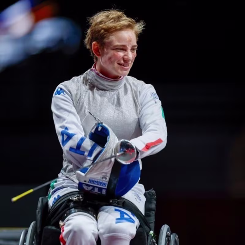 Young woman with short hair wearing a grey and blue full-body suit in a wheelchair, holding a fencing sword and smiling.
